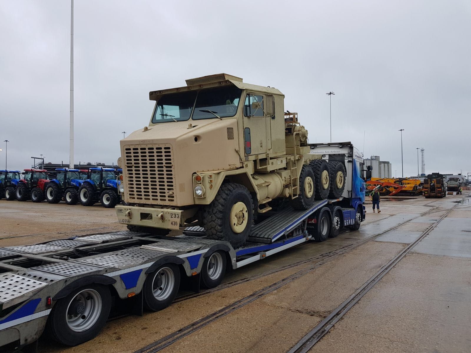 Low-loader transporter carrying a large sand-coloured military truck through a vehicle and plant yard, rows of tractors and diggers behind.