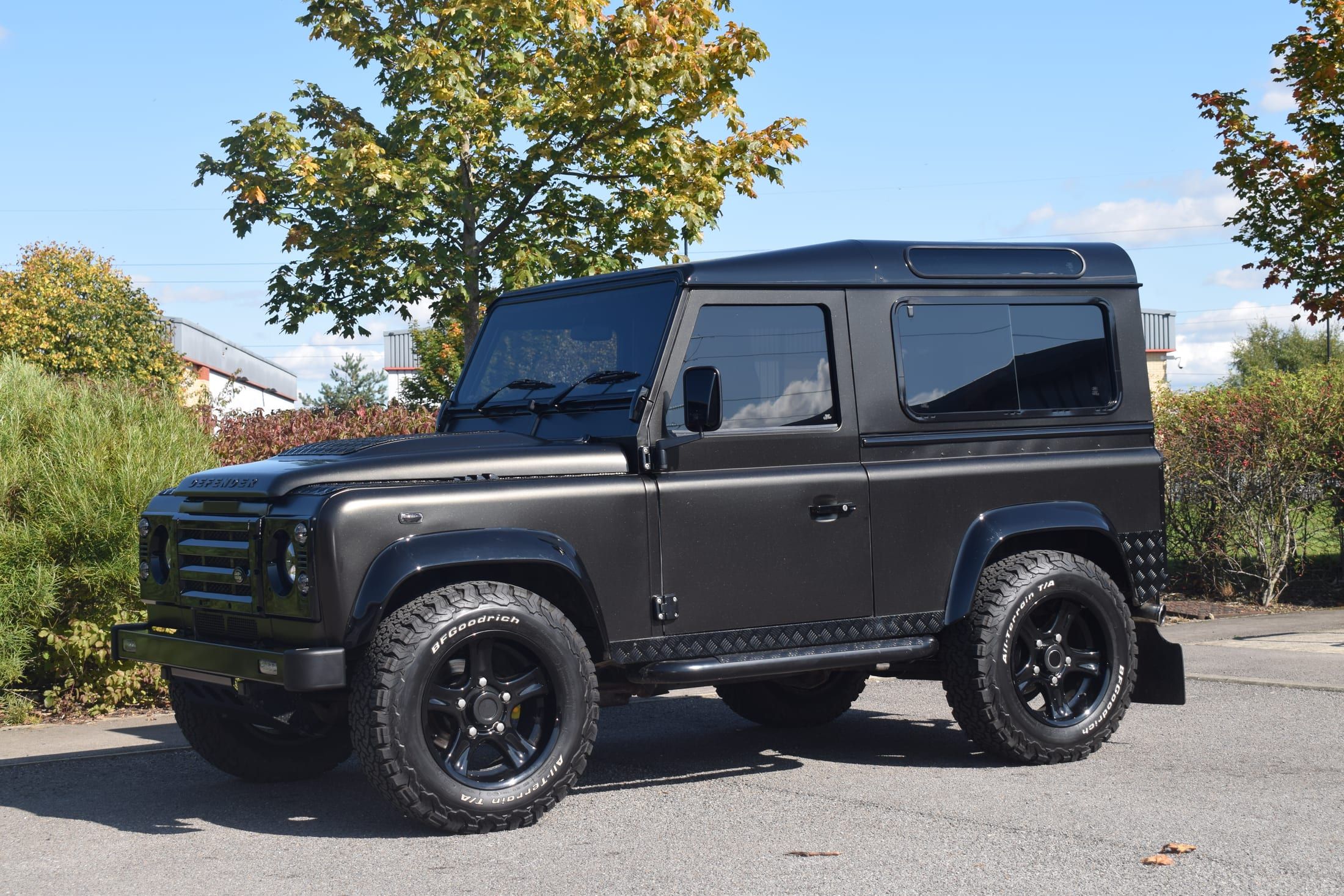 Dark graphite-grey wrapped Land Rover Defender 90 with a low-sheen finish, parked outdoors against trees and a blue sky.