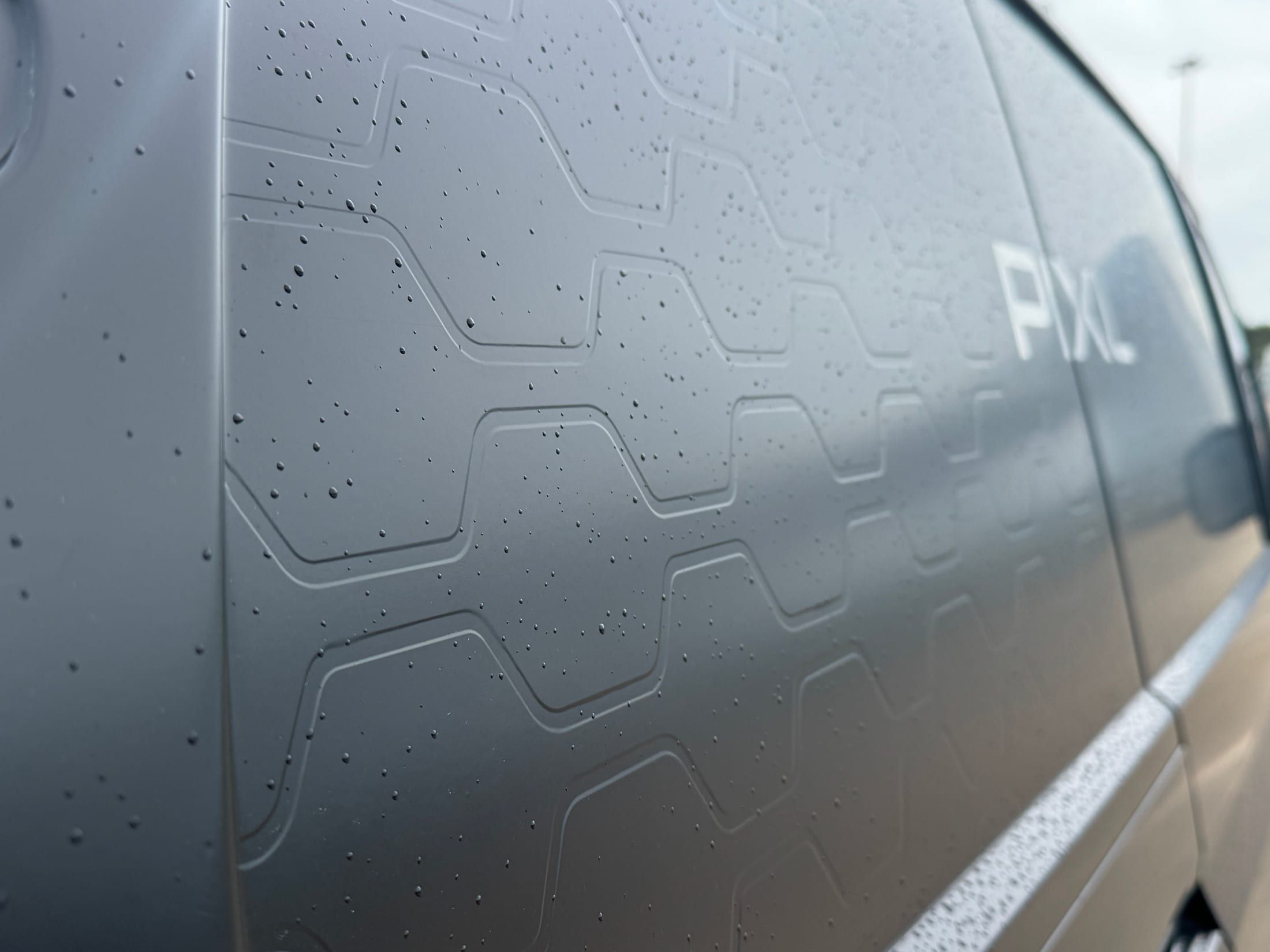 Close-up of a grey hexagonal-textured vehicle wrap on a van panel with the PIXL wordmark in white vinyl, rain beading on the surface.